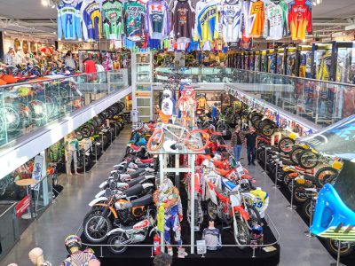 En omfattande samling av motorcyklar, särskilt motocross- och endurocyklar, visas i ett museum. Olika tröjor och utrustning hänger från taket och längs väggarna.