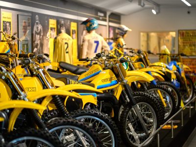 En rad med klassiska gula Suzuki motocrosscyklar med blå detaljer är uppställda i ett museum. I bakgrunden syns skärmar med information och bilder, samt tävlingströjor och hjälmar.