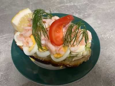 En räksmörgås med räkor, ägg, dill, en tomatskiva och en citronklyfta, serverad på en mörkgrön tallrik på ett grått underlag.