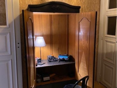 En öppen garderob av trä med svarta detaljer, fungerar som ett litet skrivbord med en lampa och föremål. En svart stol framför, ljus trävägg, hängande lampa och två vita dörrar.