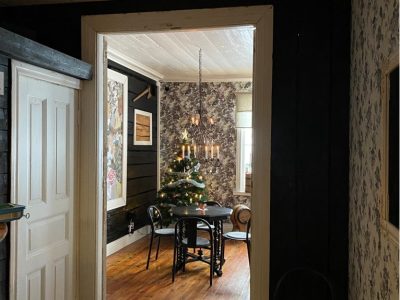 En julgran med ljus står i ett rum med mörka träväggar och ett trägolv, sett genom en dörröppning. Ett runt svart bord med stolar och en ljuskrona hänger över granen. Rummet har även tapetserade väggar och ett fönster i bakgrunden.