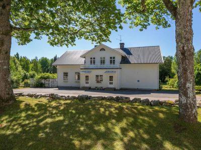 En vit tvåvåningsbyggnad med grått tak och en mindre balkong, inramad av två stora träd och en gräsmatta med stenar i förgrunden.
