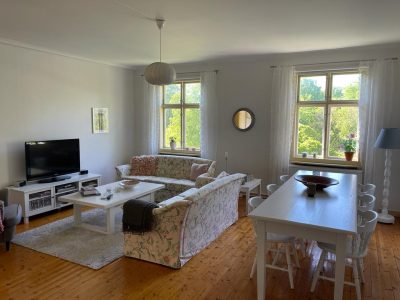 Ett ljust vardagsrum med trägolv, två soffor, ett vitt soffbord, en TV och ett matbord vid fönstret.
