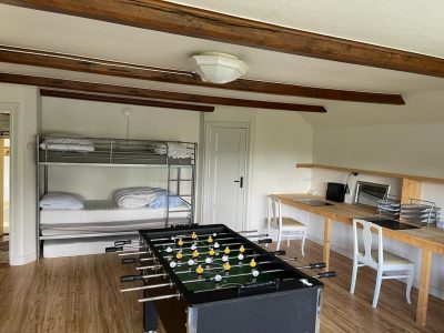 Ett rum med träbalkar i taket, en fotbollsspel i förgrunden, en våningssäng till vänster och ett långt skrivbord med stolar till höger.