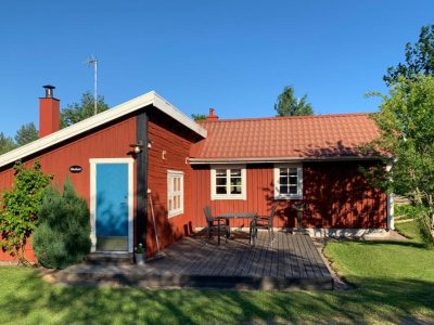 En traditionell röd svensk stuga med vita knutar, blå dörr och en träterrass med utemöbler. En grön buske står vid ingången.
