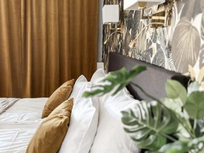 Bilden visar ett sovrum med en säng klädd i vitt och bruna kuddar. Väggen bakom sängen har en mörk tapet med bladmönster, och det finns vägglampor. I bakgrunden syns bruna gardiner.