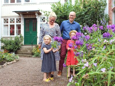 En familj på fyra, med två vuxna och två små flickor, står leende utomhus framför ett hus i Vimmerby. Flickorna håller i lila blommor.