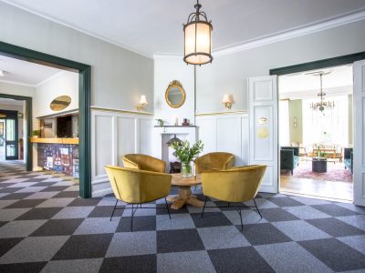 En rymlig hotellobby med schackrutigt golv i grått och svart. I mitten finns en grupp med tre senapsgula sammetsfåtöljer runt ett litet träbord. En öppen dörr till vänster leder till en reception med en färgglad disk. En öppen dörr till höger leder till ett vardagsrum med grön inredning. Ett fönster och en öppen spis med en spegel ovanför syns i bakgrunden.