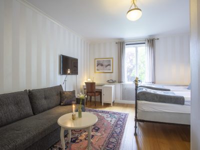 Ett hotellrum med en grå soffa i förgrunden, ett vitt soffbord med levande ljus, och en mönstrad matta. Rummet har en tv på väggen, ett skrivbord, och en säng med ett smidesjärn i fotänden. Ett fönster med ljusa gardiner släpper in dagsljus.