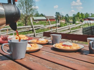En hand häller kaffe från en termos i en grå mugg på ett rustikt träbord. På bordet står även tallrikar med våfflor toppade med grädde och sylt, samt fler kaffekoppar. I bakgrunden syns ett pittoreskt landskap med röda stugor och ett trästaket.