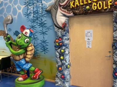 En stor skulptur av en sköldpadda med golfklubba står bredvid en dörr med en skylt märkt "Kalles Golf", flankerad av en haj och med en undervattensmural på väggen.