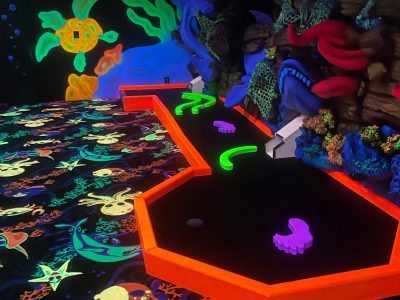 En bana för minigolf i neonfärger med ett undervattenstema. Golvet har självlysande bilder av havsdjur, och banans kanter lyser starkt orange. I bakgrunden finns en väggmålning med korallrev och havsliv som också lyser i neon.
