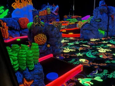 En självlysande minigolfbana med undervattenstema, där banorna lyser i neonorange. Golvet är dekorerat med självlysande havsdjur, och omgivningen har skulpturer föreställande koraller och klippor som lyser i starka färger.