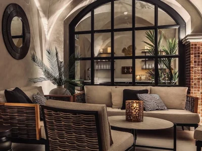 En elegant lounge med bekväma sittmöbler, en stor välvd fönstervägg med utsikt över ett annat rum, och detaljer som växter och en kopparmosaikvägg.
