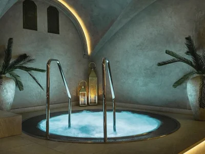 En jacuzzi med bubblande, blått upplyst vatten och silverfärgade räcken. Rummet har bågformade valv i taket med dold belysning, grå väggar och är dekorerat med gröna palmer i stora krukor samt gyllene lyktor.