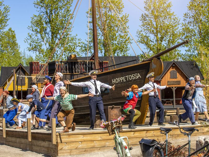 En scenbild med skådespelare i färgglada kostymer från föreställningen Pippi Långstrump, dansar och sjunger framför ett stort träskepp märkt "HOPPE TOSSA". Föreställningen äger rum utomhus på en solig dag, med träd och hus i bakgrunden.