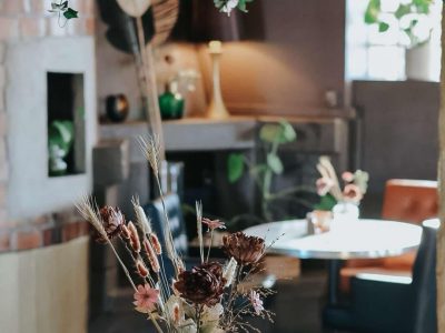 En vas med torkade blommor på ett träbord, med en takdekoration av grönska och en mysig restaurangmiljö i bakgrunden.