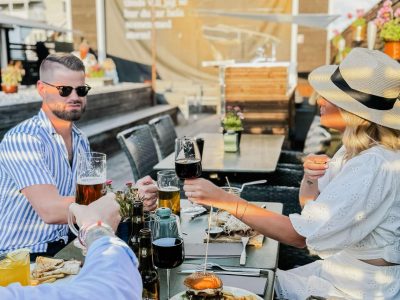 En grupp vänner skålar med drycker vid ett bord på en uteservering. De har mat, inklusive hamburgare med pommes frites, framför sig. Bakom dem syns andra sittplatser, parasoller och en byggnad.