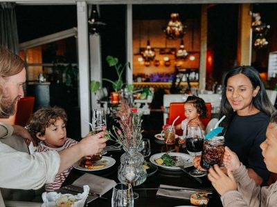 En familj på fyra sitter vid ett bord i en restaurang och skålar med sina drycker. De har olika rätter framför sig, och bakgrunden är suddig men visar en del av restauranginteriören med lampor och växter.