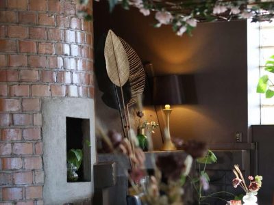 En närbild på en tegelvägg med en guldfärgad panel i en restaurangmiljö. Ett utskuret hål i väggen visar en liten växt, och i bakgrunden syns en lampa, ett bord med stolar och en takdekoration med grönska.