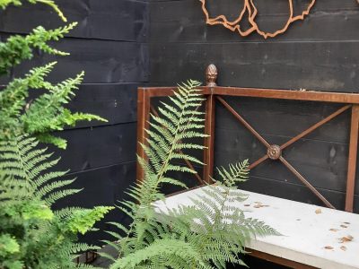 Trädgårdsbänk med järnram och vit sits, mot en svart trävägg med metallblommor, omgiven av ormbunkar.