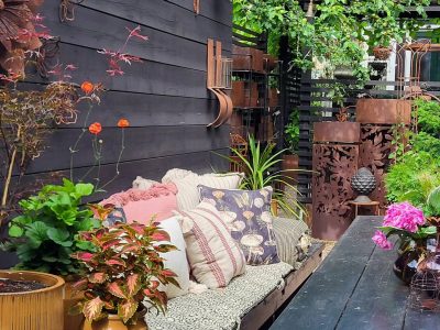 En mysig utomhusplats med en soffa fylld med kuddar, omgiven av växter och dekorativa metallföremål, intill en mörk trävägg.