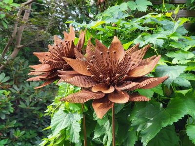 Två rostfärgade metallblommor står bland grönskande växter, en del av en trädgårdsdekoration i Vimmerby.