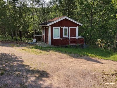 Röd stuga med veranda vid en grusplan och omgiven av lövskog.