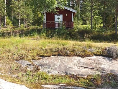 En liten röd stuga med vit dörr och veranda, omgiven av tallskog och gräs, med berghällar i förgrunden.