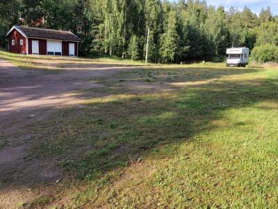 En campingplats med en röd stuga till vänster och en husbil till höger, omgiven av skog och gräs.