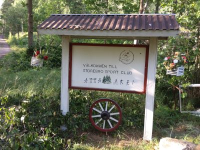 En välkomstskylt för Storebro Sport Club med en gammal vagnshjul framför, omgiven av grönska.