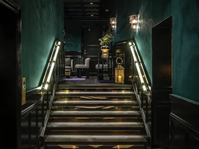 En upplyst trappa med mörkgröna väggar och svarta träpaneler, som leder upp till en elegant hall med en lampa och en dekorativ lykta.