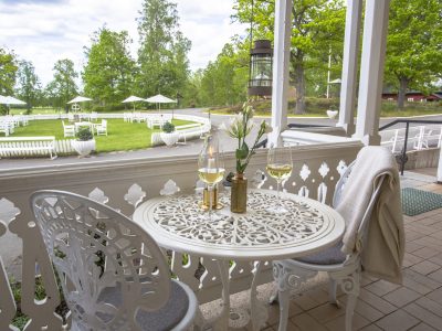 En vit uteplats med ett bord och två stolar, med två glas vitt vin och blommor. I bakgrunden syns en grön gräsmatta med fler vita utemöbler under parasoller.