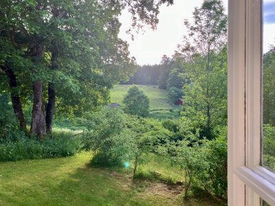 Utsikt från ett fönster över en lummig gräsmatta som övergår i en grönskande dalgång med skog och en liten röd stuga i fjärran under en ljus himmel.