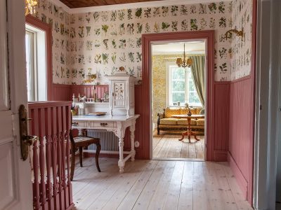 En rumsvy med ljusa trägolv och tapeter med botaniska motiv ovanför rosa träpaneler. I mitten finns ett vitt skrivbord med en vit byrå och en stol. Till höger leder en öppen dörröppning in till ett annat rum med en soffa och ett fönster. Till vänster syns en del av en vit dörr med mässingshandtag och ett rosa räcke.