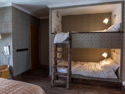 Ett mysigt sovrum med inbyggda våningssängar, var och en med en läslampa och mönstrad tapet. En trästege leder upp till den övre bädden. Rummet har trägolv och mönstrad tapet på väggarna. En dörr syns i bakgrunden.