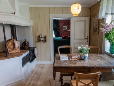 En lantlig köksmiljö med ljusa trägolv, beige panelväggar och ett stort vitt murspis med en svart vedspis. Ett träbord med stolar står i rummets mitt. Genom en öppen dörr i bakgrunden skymtar ett sovrum med en mörkgrön säng och röd tapet. Ett fönster till höger släpper in ljus och är dekorerat med en vas med lila blommor.