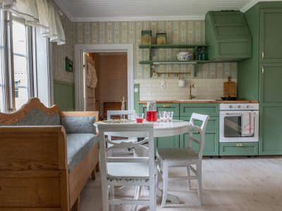 Ett lantligt kök med ljusa trägolv och gröna skåp och snickerier. En vit rund matbord med två vita stolar står i rummets mitt. Till vänster finns en träsoffa framför ett stort fönster. Köket har en vit kakelvägg över diskbänken och öppna hyllor. En dörröppning i bakgrunden leder till ett annat rum.