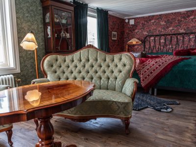 Ett ombonat rum med en grön stoppad soffa i förgrunden, ett träbord framför den, och en säng med rött överkast i bakgrunden. Väggarna har mönstrade tapeter och rummet är inrett med klassiska möbler och lampor.