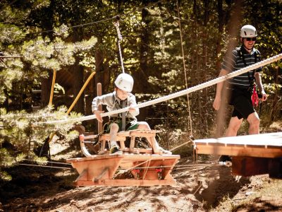 En vuxen och ett barn med hjälmar och säkerhetsselar deltar i ett höghöjdsbana i en skog. Barnet sitter på en träplattform som glider längs ett rep, medan den vuxne går bredvid.